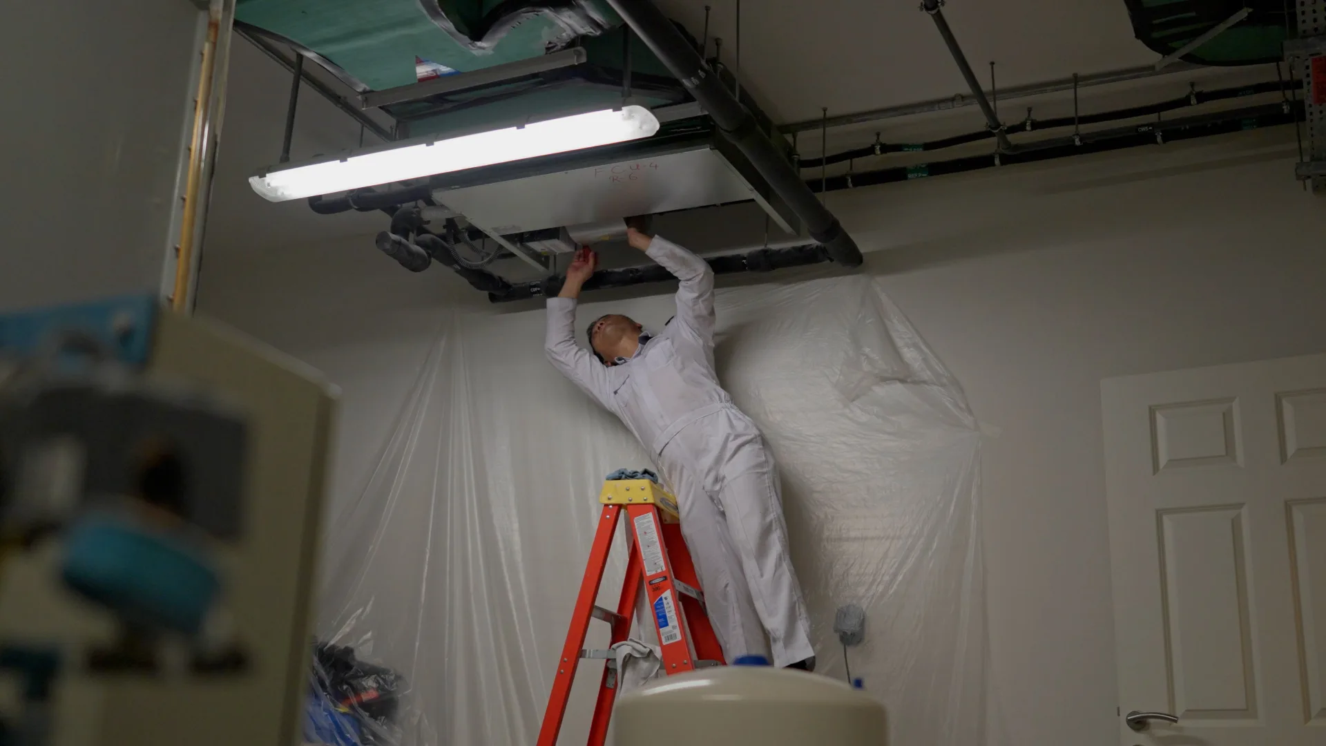 A Saniservice technician servicing a ceiling fan-coil unit from a ladder